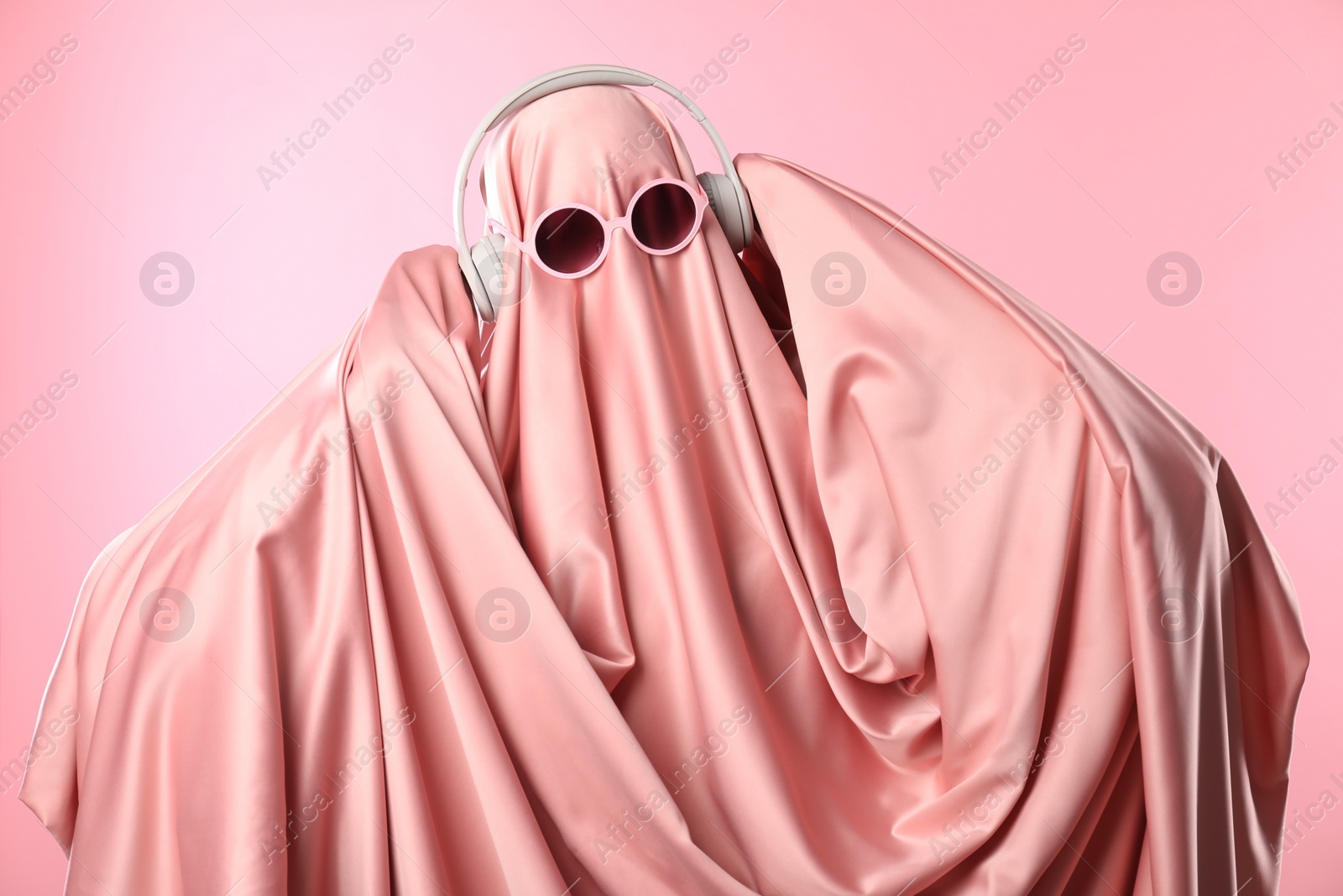 Photo of Glamorous ghost. Woman in sheet with sunglasses and headphones on pink background