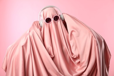 Photo of Glamorous ghost. Woman in sheet with sunglasses and headphones on pink background