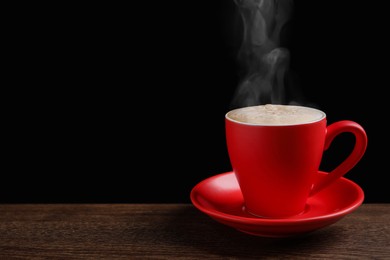 Red cup with hot steaming coffee on wooden table against black background, space for text