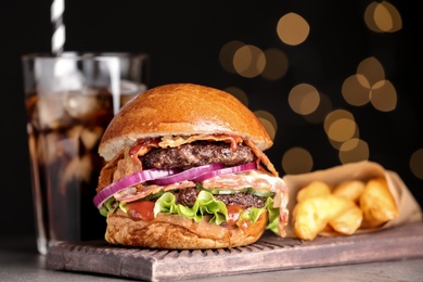 Board with burger and cola on table against blurred lights