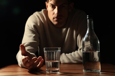 Photo of Addicted man with alcoholic drink at wooden table against black background, focus on glass