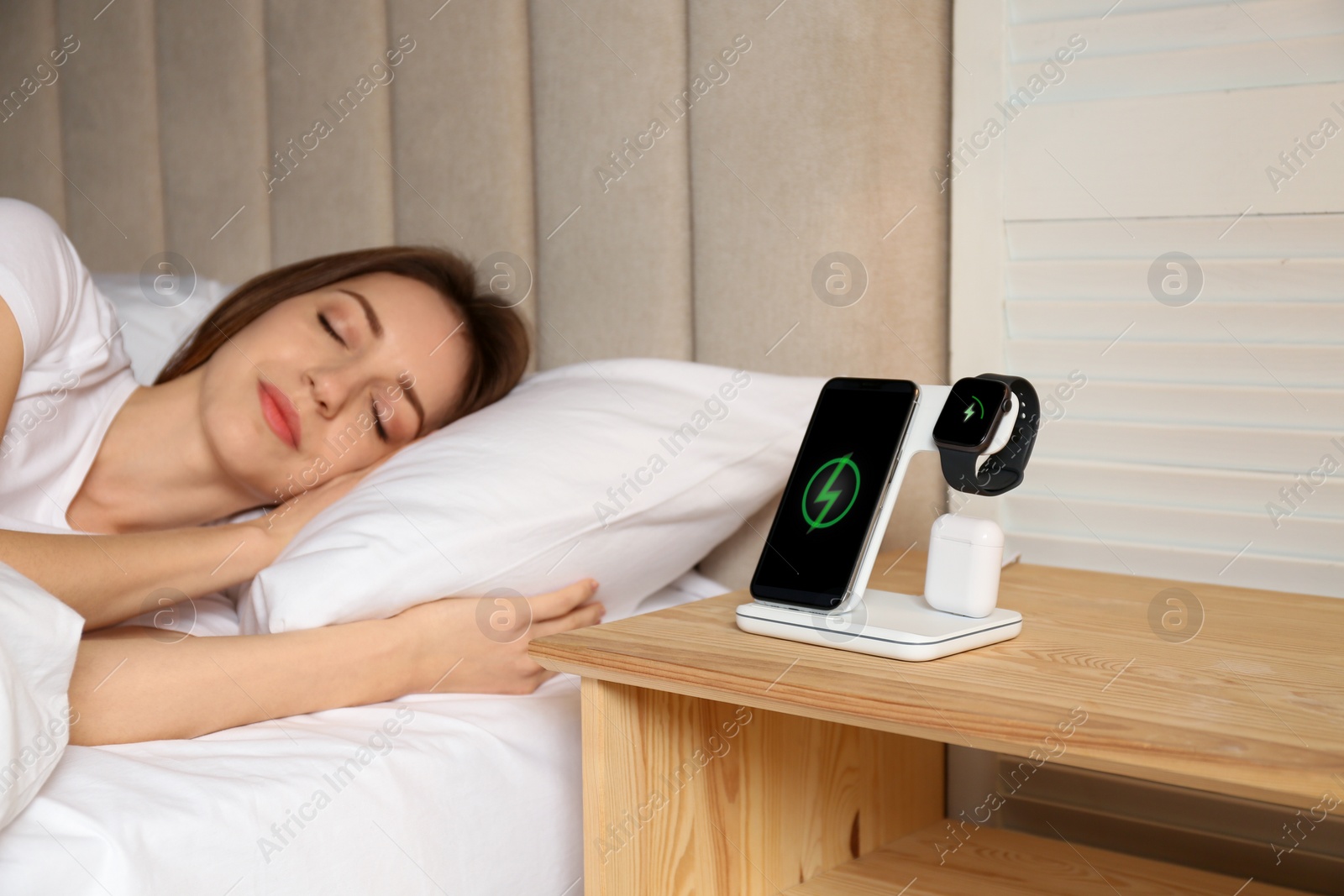 Photo of Smartphone, watch, earphones charging on wireless pad and sleeping woman in room