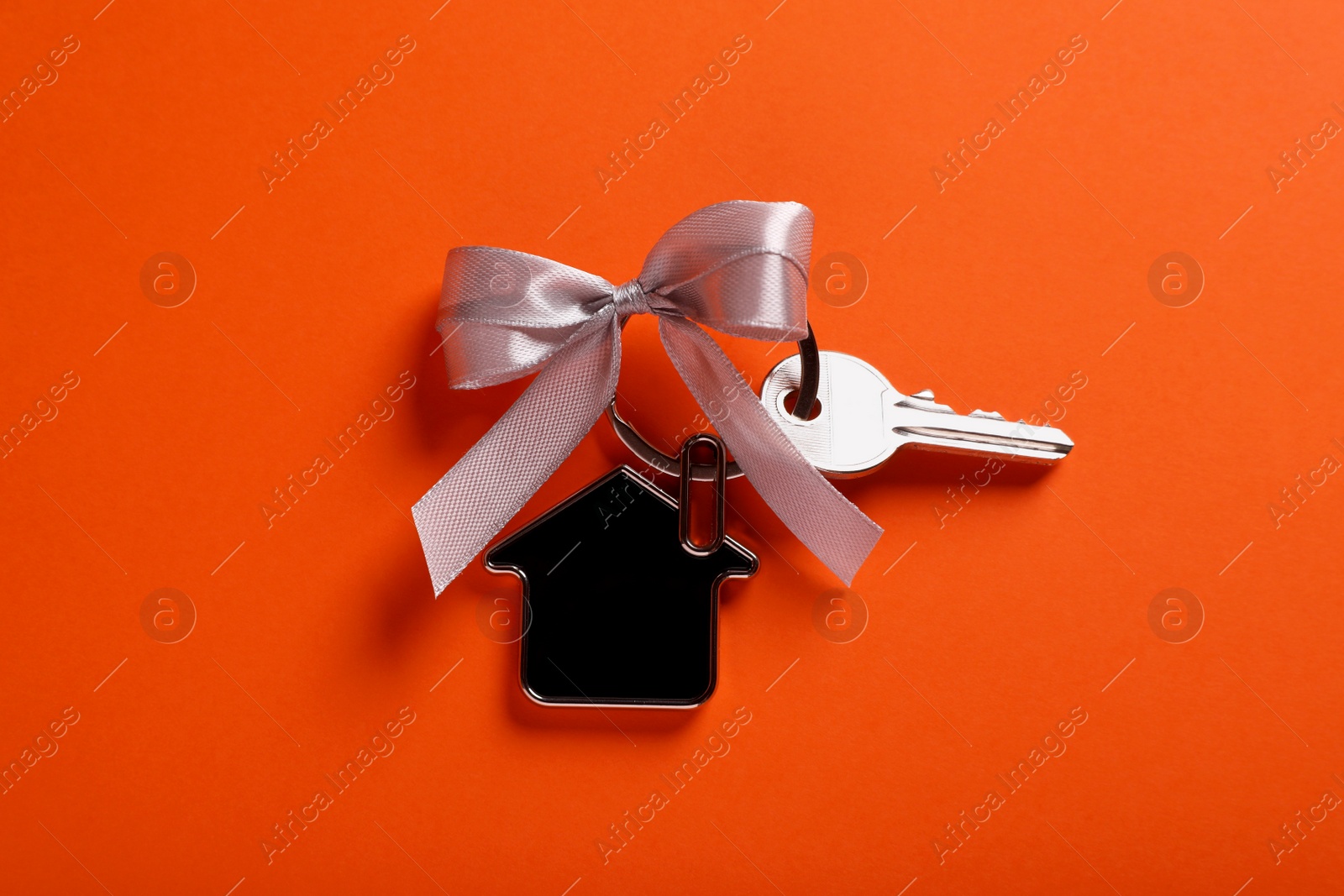 Photo of Key with trinket in shape of house and grey bow on orange background, top view. Housewarming party