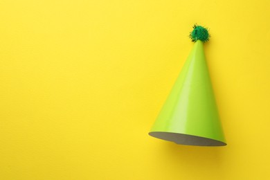 One green party hat with pompom on yellow background, top view. Space for text