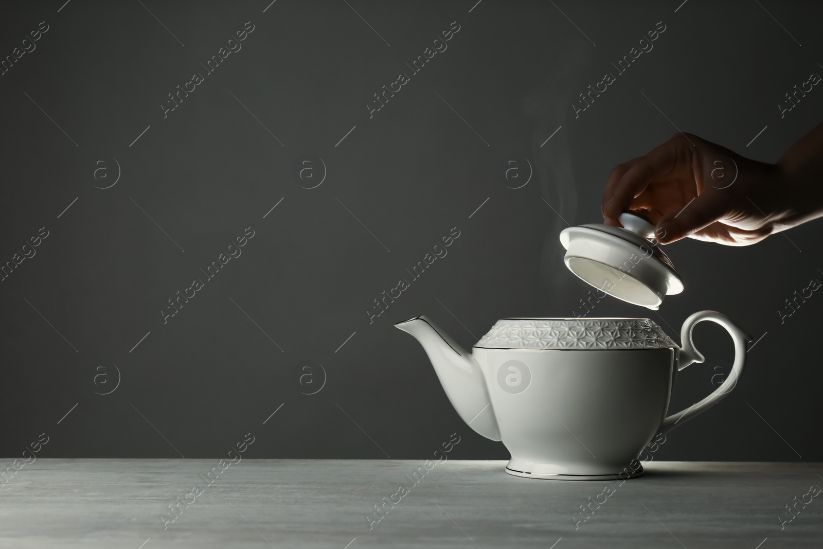 Photo of Woman removing lid from ceramic teapot at table against grey background, closeup. Space for text