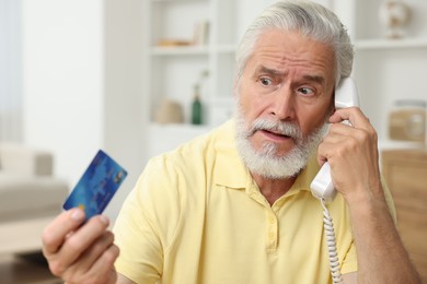 Photo of Confused senior man with credit card talking on phone at home. Be careful - fraud