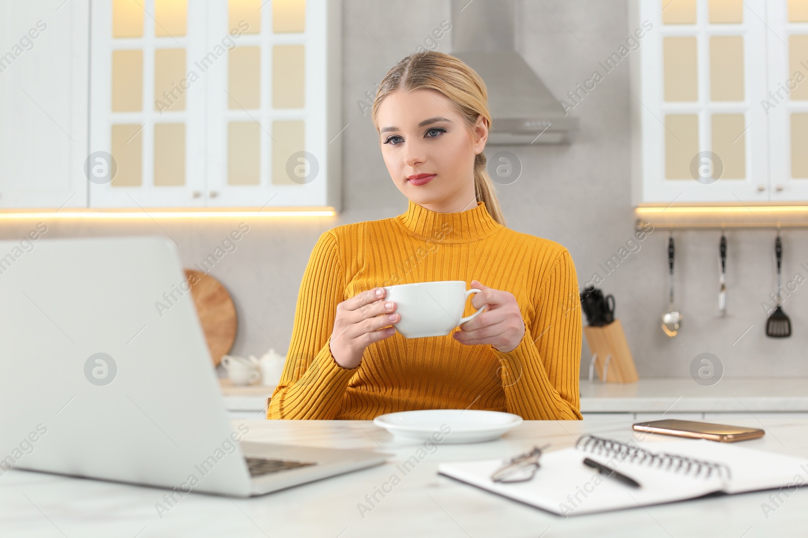 Photo of Home workplace. Woman with cup of hot drink looking at laptop at marble desk in kitchen