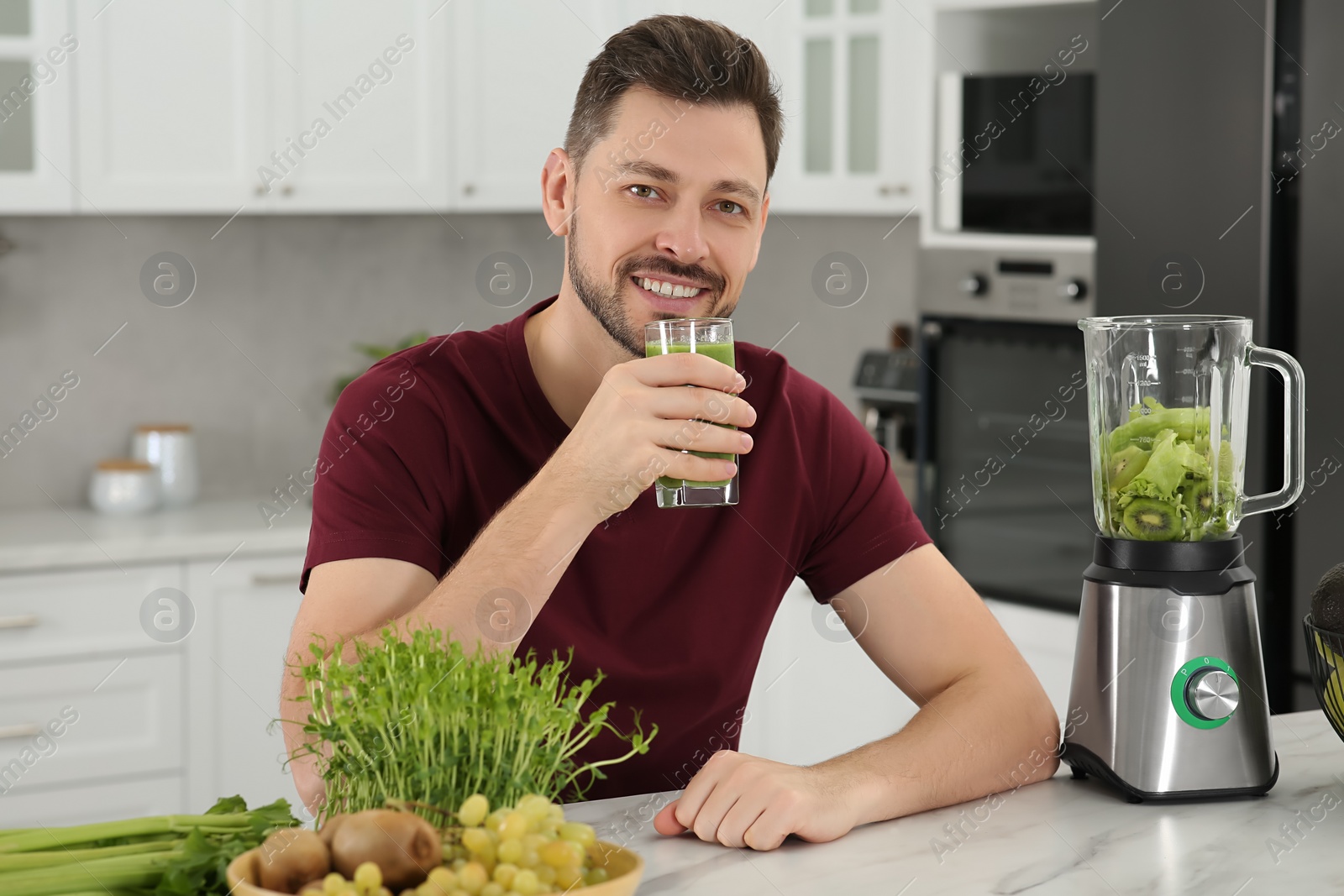 Photo of Happy man drinking delicious smoothie at white marble table in kitchen