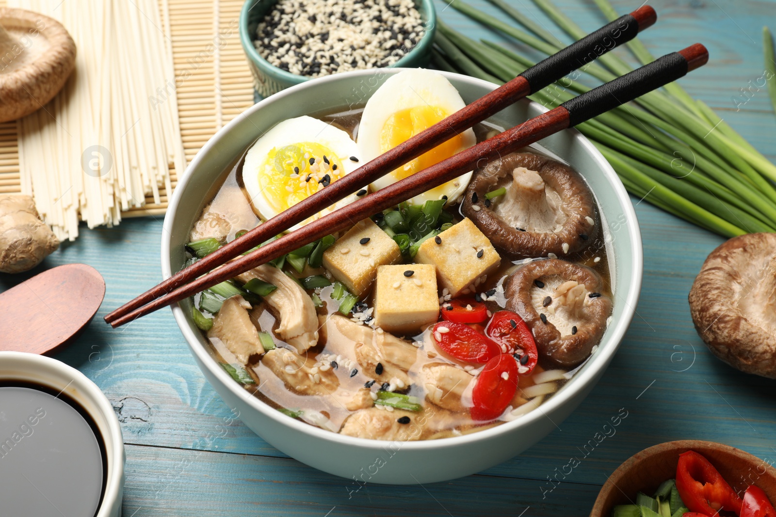 Photo of Bowl of delicious ramen and ingredients on light blue wooden table. Noodle soup