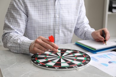 Business targeting concept. Man with dart aiming at dartboard at gray marble table indoors, closeup