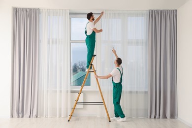 Photo of Workers in uniform hanging window curtain indoors