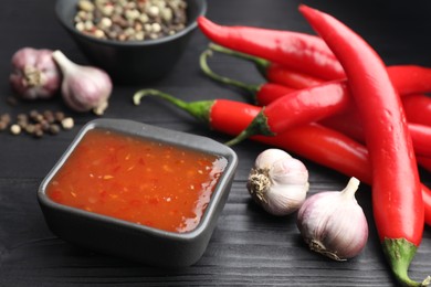 Photo of Spicy chili sauce, garlic, peppers and peppercorns on black wooden table, closeup