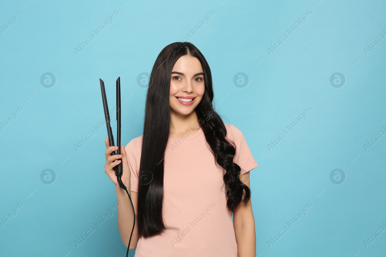 Photo of Beautiful happy woman with hair iron on light blue background