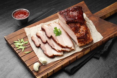 Photo of Pieces of baked pork belly served with sauce and parsley on black textured table