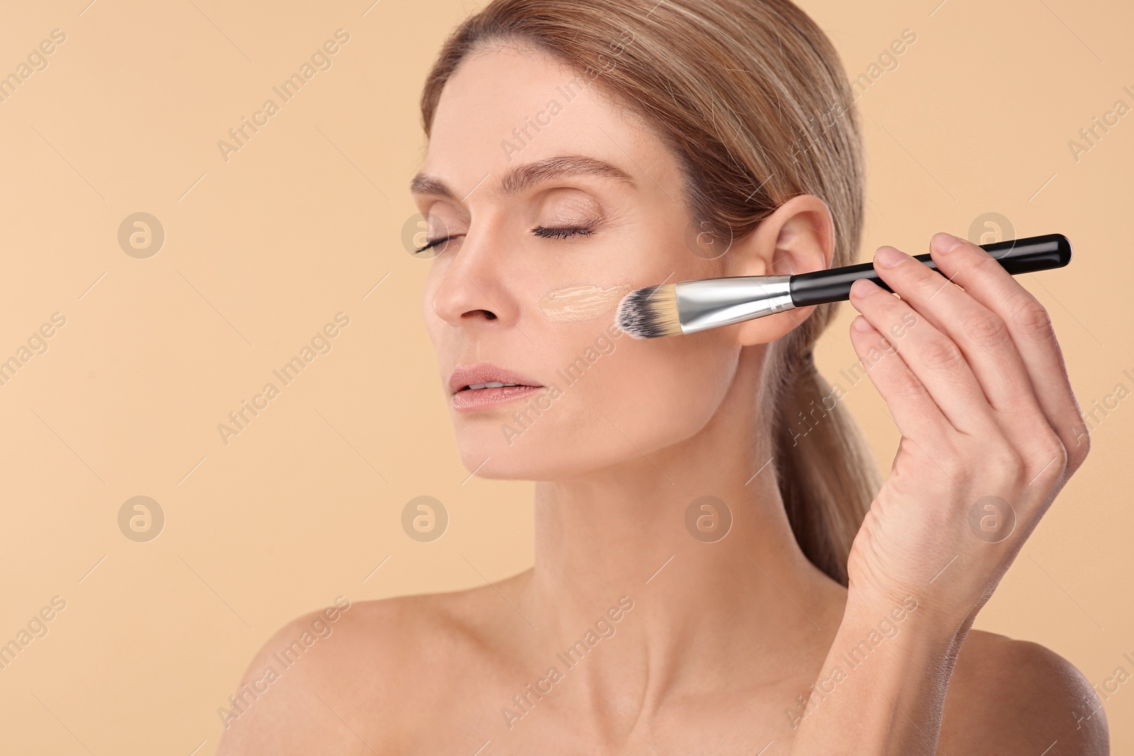 Photo of Woman applying foundation on face with brush against beige background