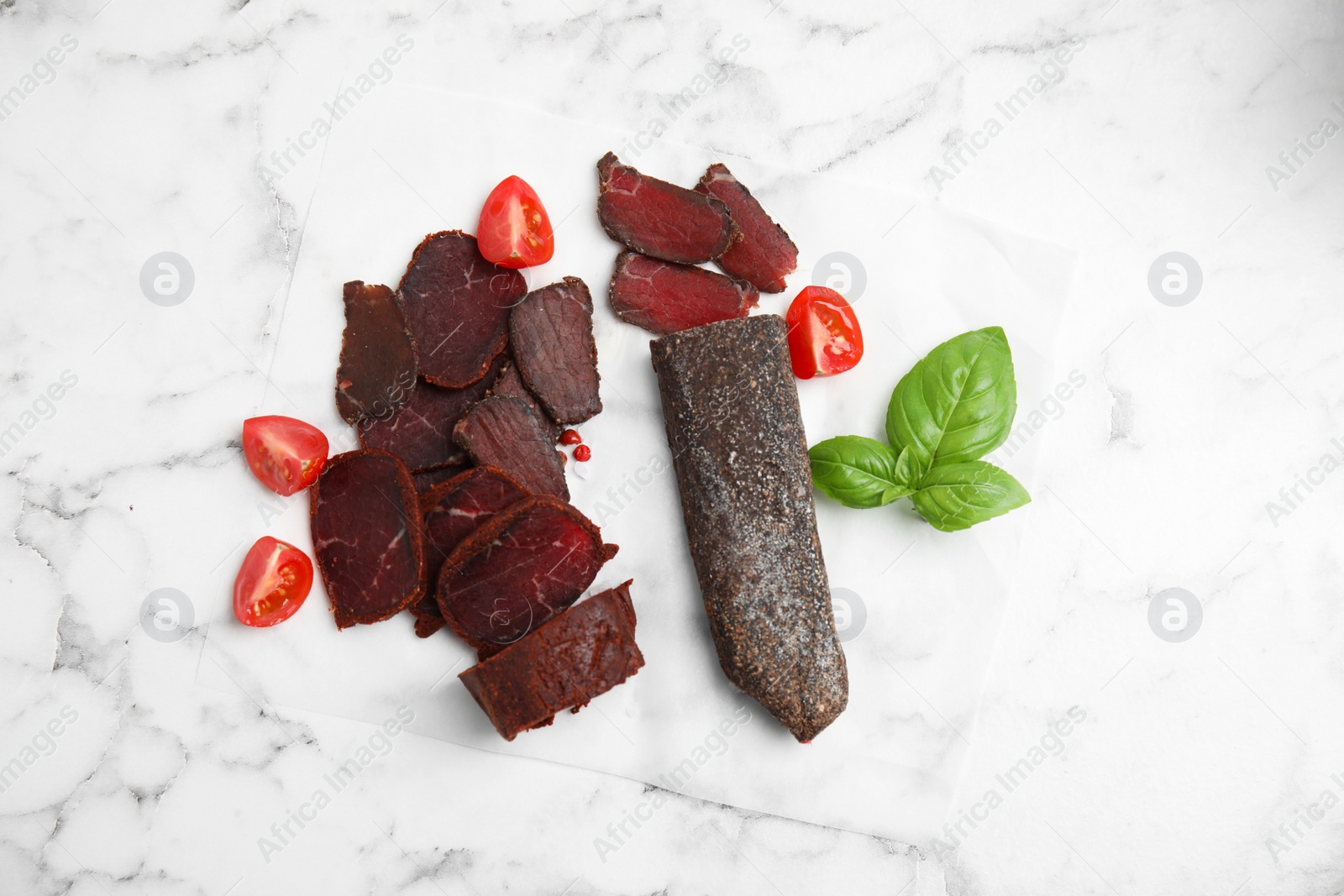 Photo of Delicious dry-cured beef basturma with basil and tomatoes on white marble table, flat lay