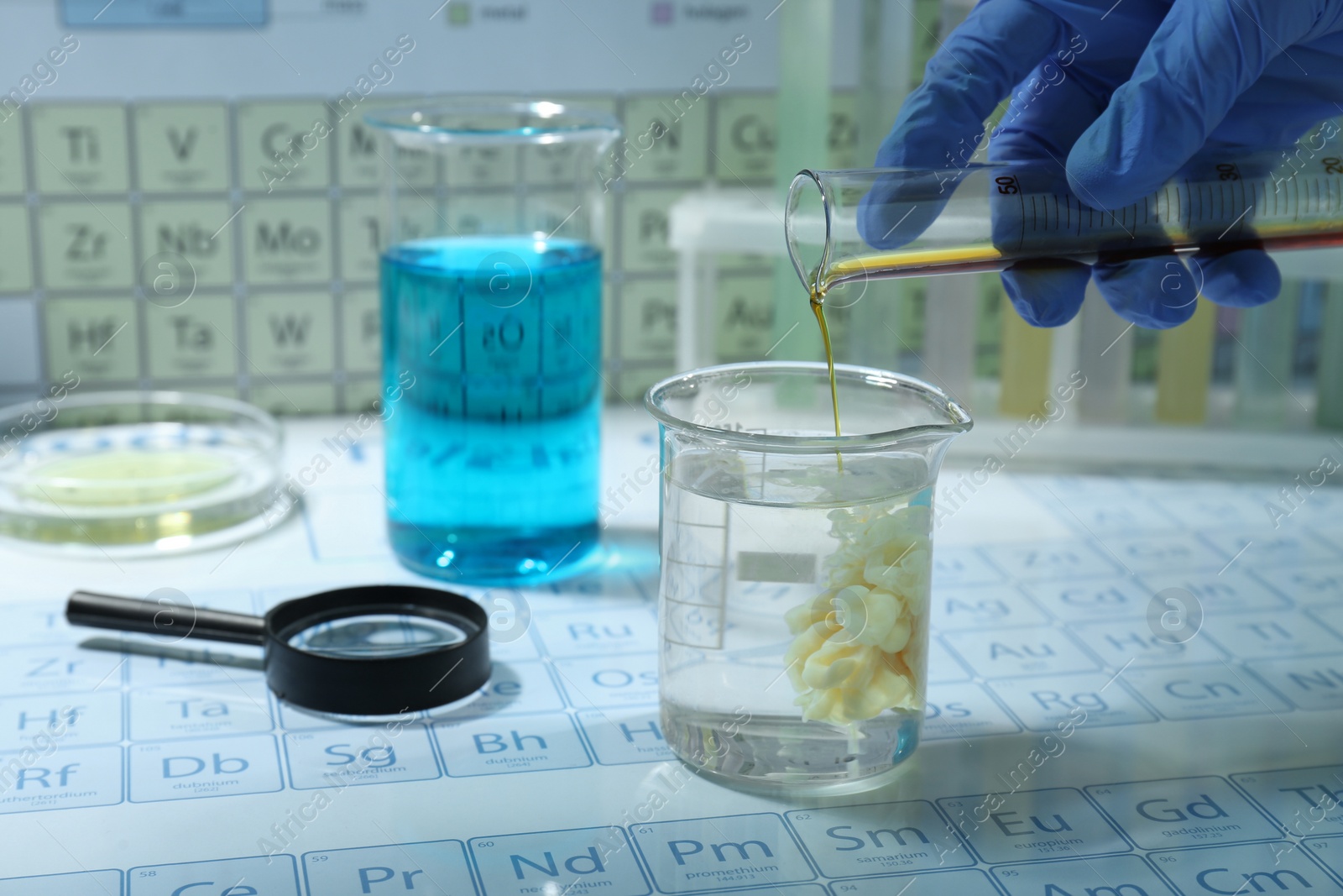 Photo of Scientist pouring liquid from test tube into beaker, different laboratory glassware and magnifying glass on periodic table of chemical elements, closeup