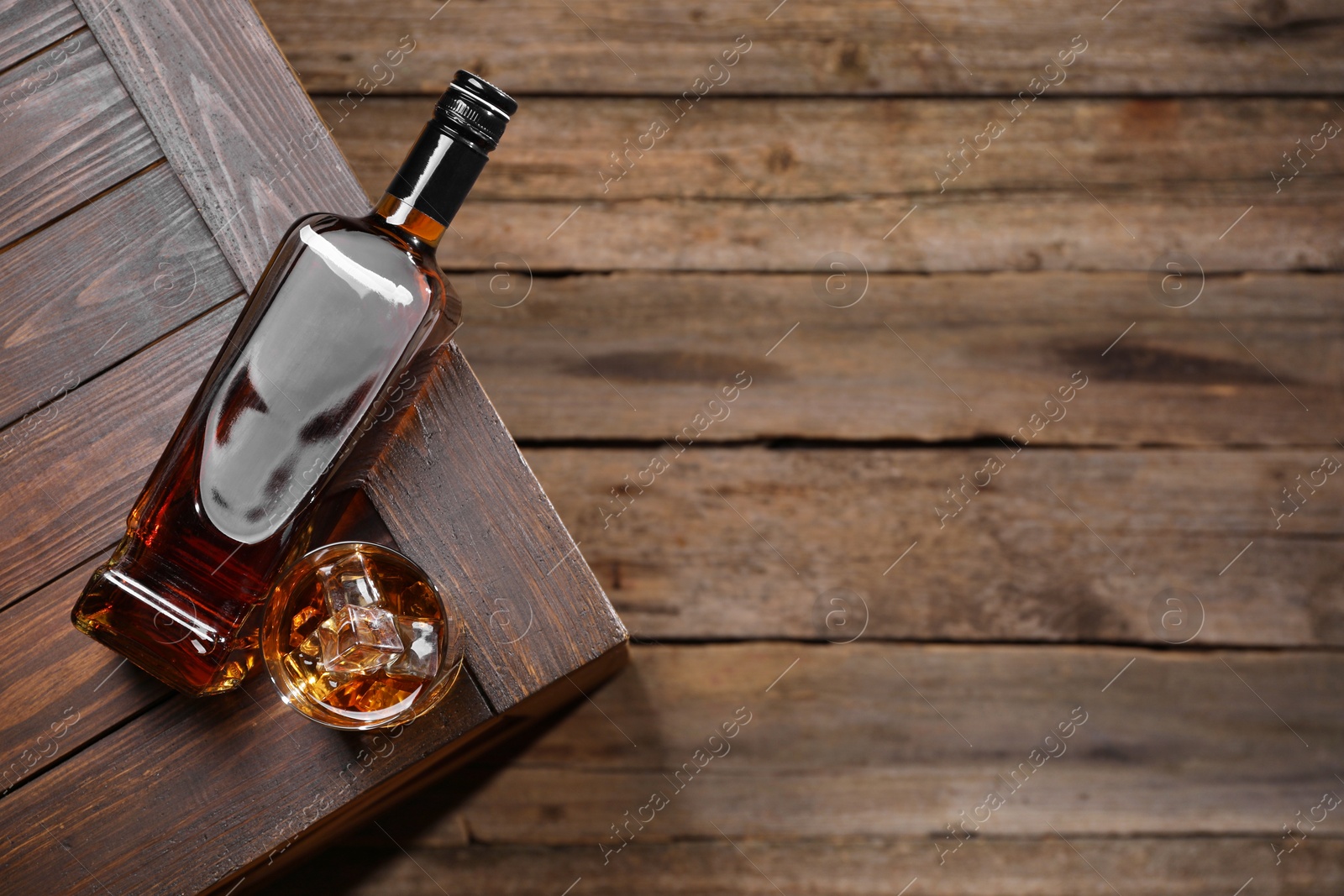 Photo of Whiskey with ice cubes in glass and bottle on wooden crate, top view. Space for text