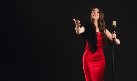 Beautiful young woman in stylish dress with microphone singing on black background