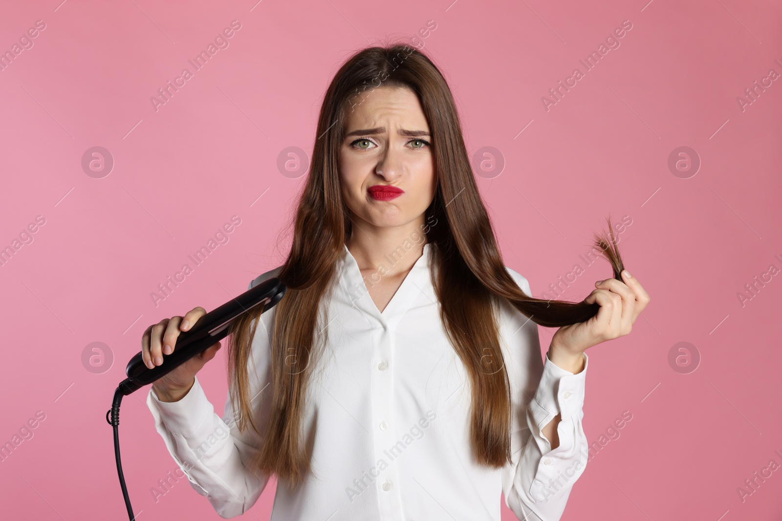 Photo of Upset young woman with flattening iron on light pink background. Hair damage