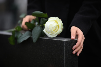 Photo of Woman holding white rose near black granite tombstone outdoors, closeup. Funeral ceremony