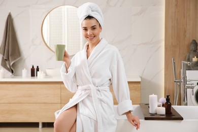 Beautiful young woman with cup sitting on edge of tub in bathroom