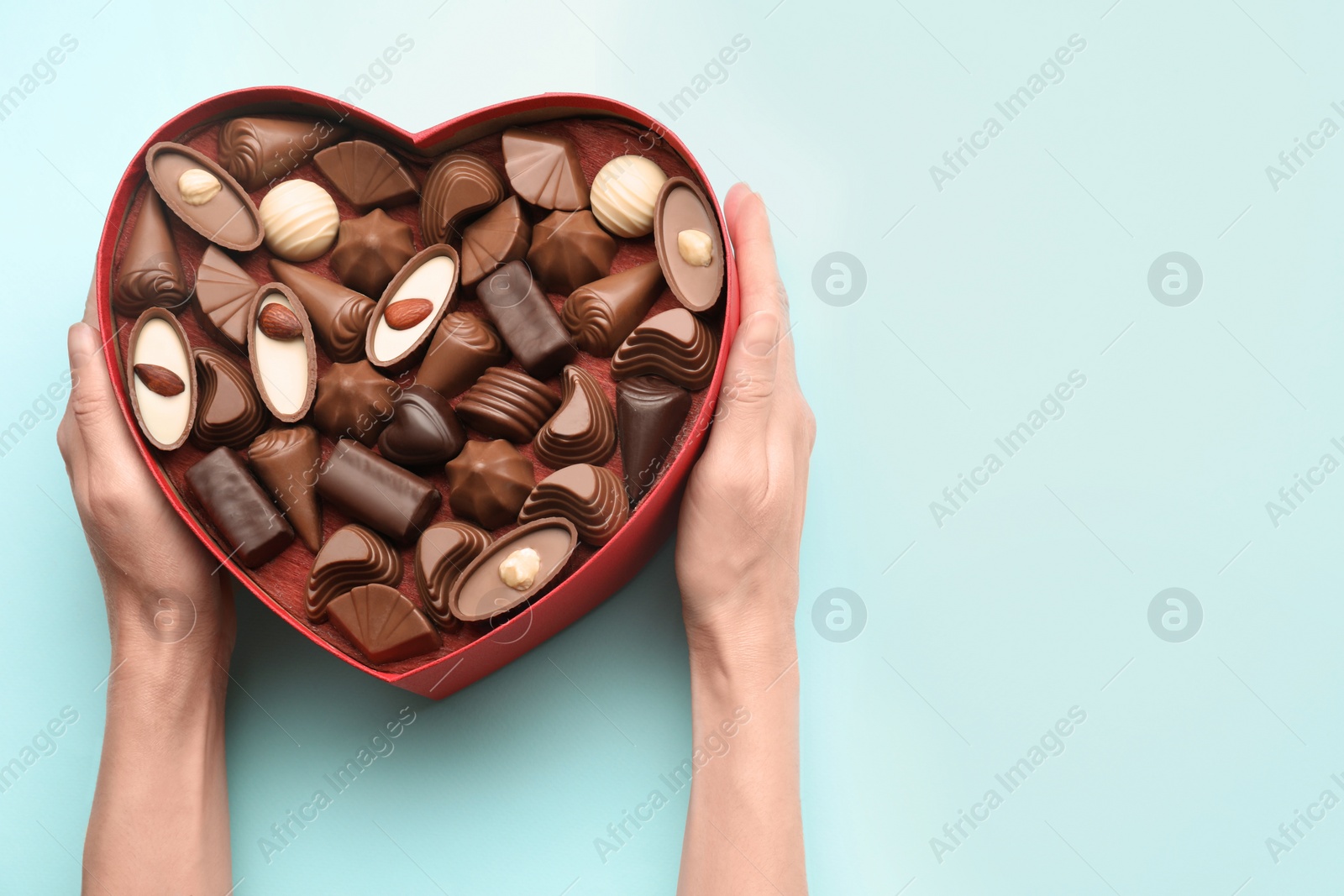 Photo of Woman holding heart shaped box with delicious chocolate candies on light blue background, top view. Space for text