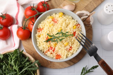 Photo of Tasty couscous with pepper, corn and arugula served on white tiled table, flat lay