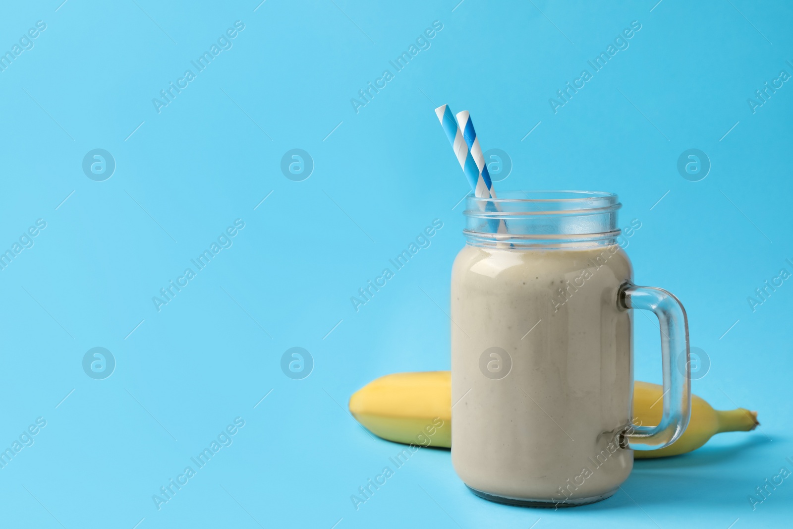 Photo of Mason jar of tasty banana smoothie with straws and fresh fruit on light blue background. Space for text