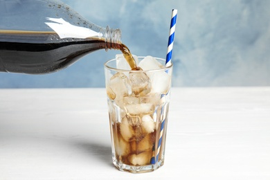 Pouring refreshing soda drink into glass with ice cubes on white wooden table against blue background