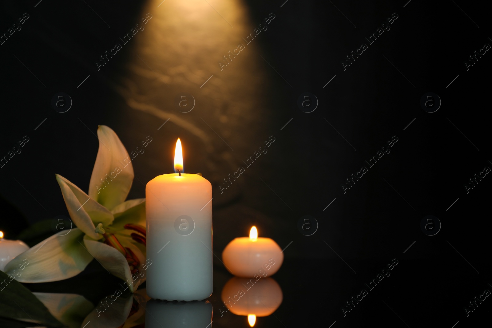 Photo of White lily and burning candles on black mirror surface in darkness, space for text. Funeral symbols