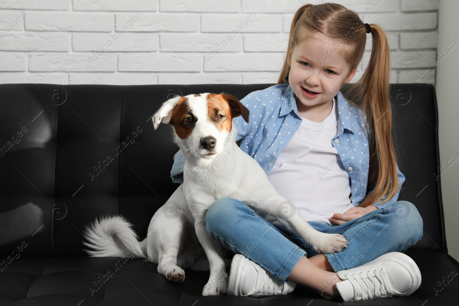 Photo of Cute little girl with her dog on sofa indoors. Childhood pet