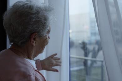 Photo of Elderly woman looking out of window indoors, space for text. Loneliness concept