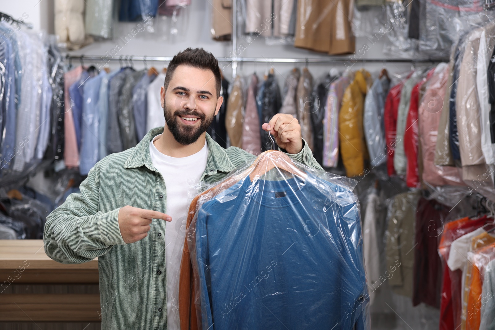 Photo of Dry-cleaning service. Happy man holding hangers with clothes in plastic bags indoors