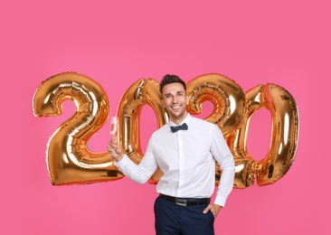 Photo of Happy young man with glass of champagne near golden 2020 balloons on pink background. New Year celebration