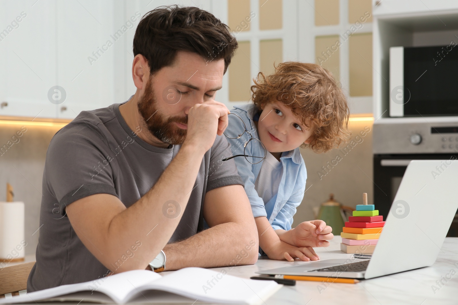 Photo of Little boy bothering father while he working remotely at home. Man with laptop and his child at desk in kitchen