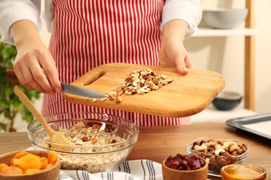 Photo of Making granola. Woman adding nuts into bowl with oat flakes at table in kitchen, closeup