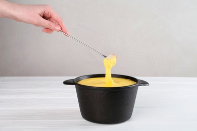 Woman dipping piece of bread into fondue pot with tasty melted cheese at white wooden table against gray background, closeup