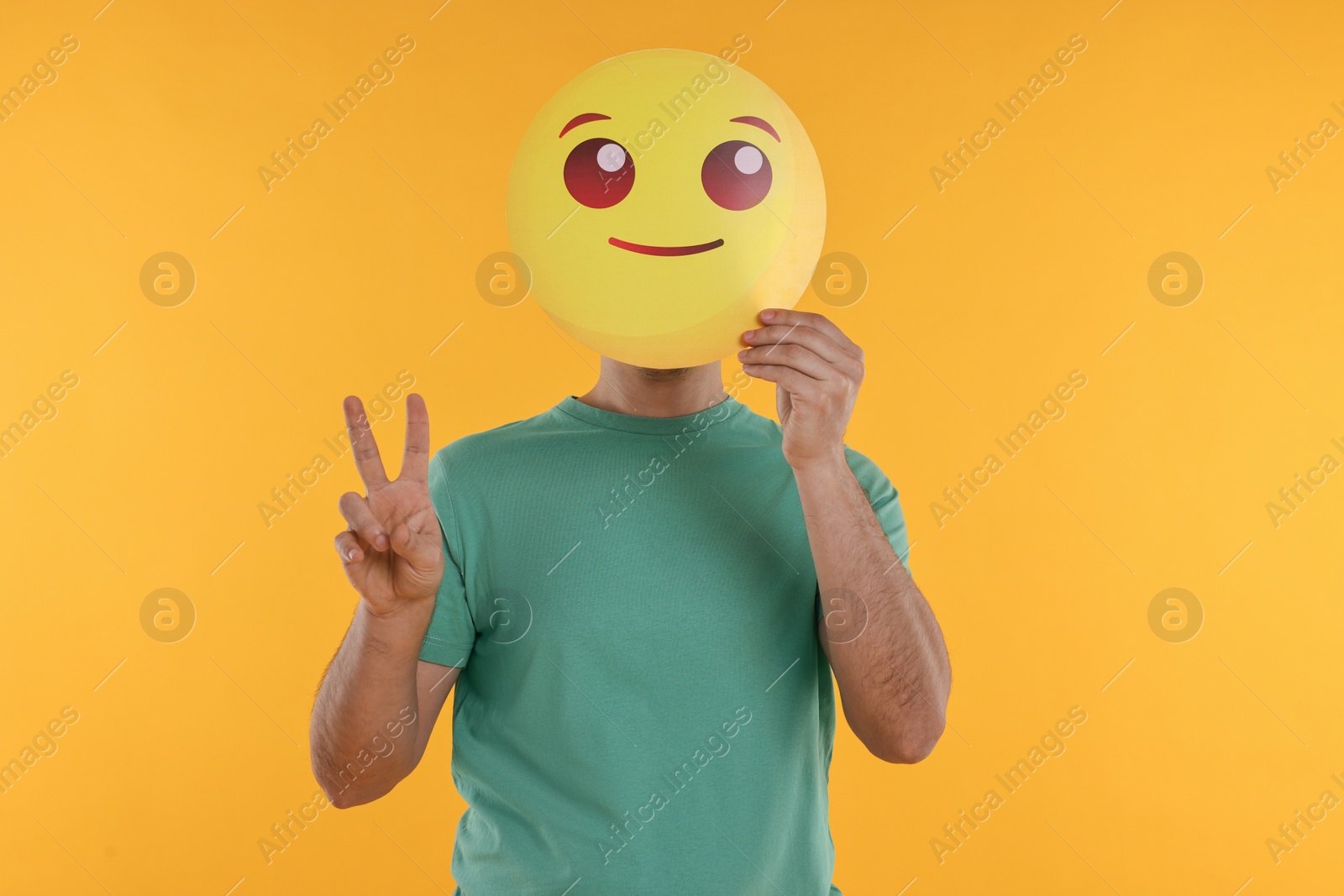 Photo of Man covering face with smiling emoticon and showing peace sign on yellow background