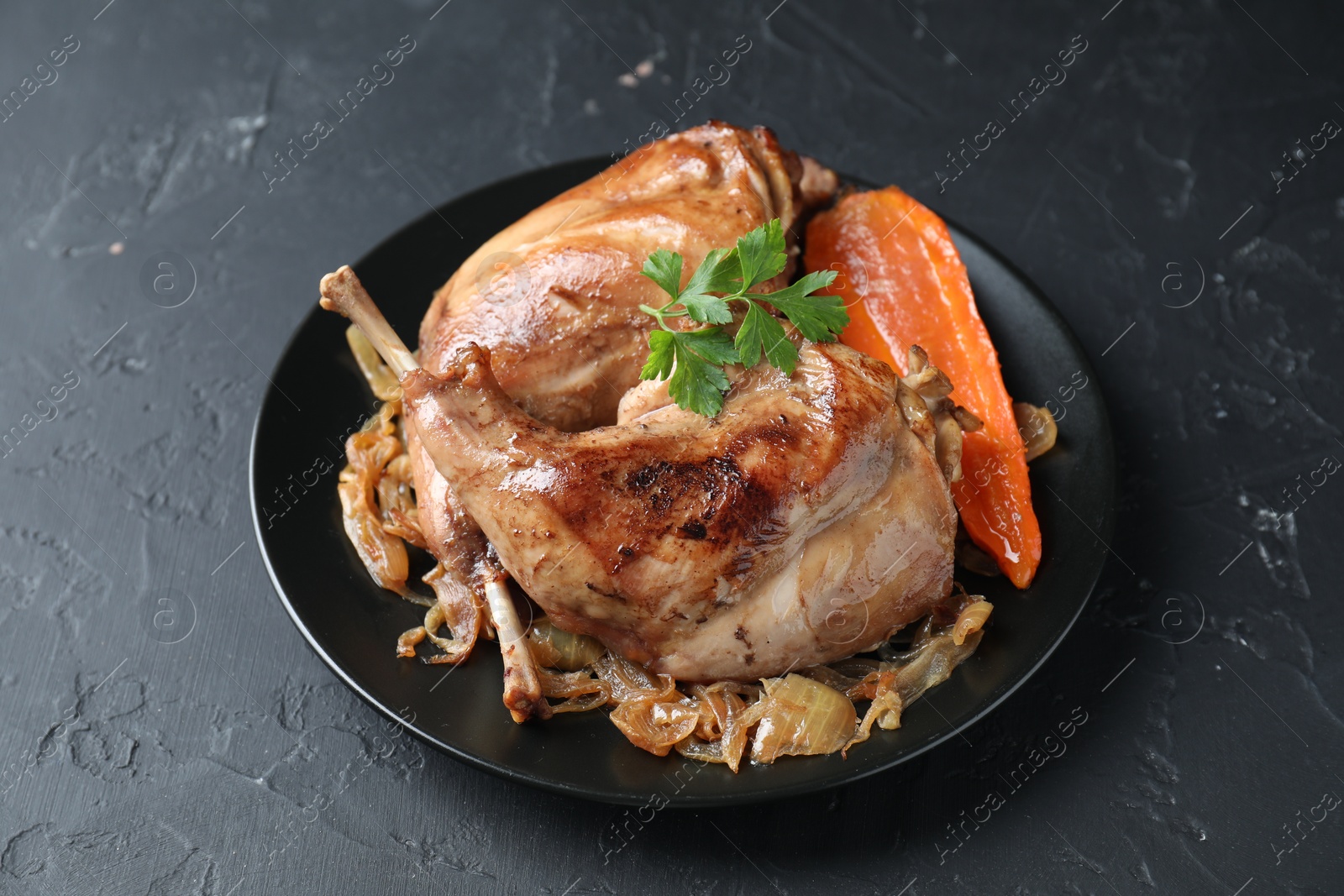 Photo of Tasty cooked rabbit meat with vegetables and parsley on black table