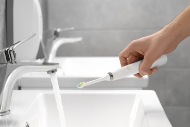 Man holding electric toothbrush near flowing water above sink in bathroom, closeup
