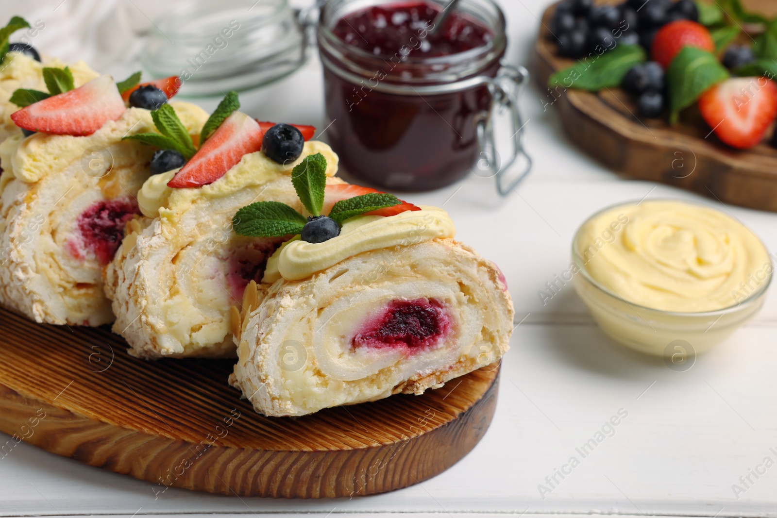 Photo of Pieces of tasty meringue roll with jam, cream, strawberry, blueberry and mint on white wooden table, closeup