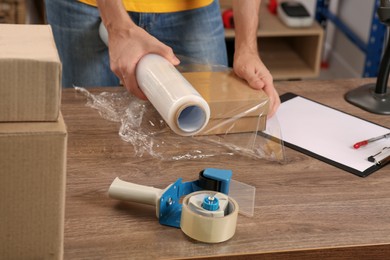 Photo of Post office worker wrapping parcel in stretch film at counter indoors, closeup