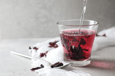 Photo of Making aromatic hibiscus tea. Pouring water into glass with dried roselle calyces and spoon on light table, space for text