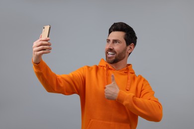Photo of Smiling man taking selfie with smartphone and showing thumbs up on grey background