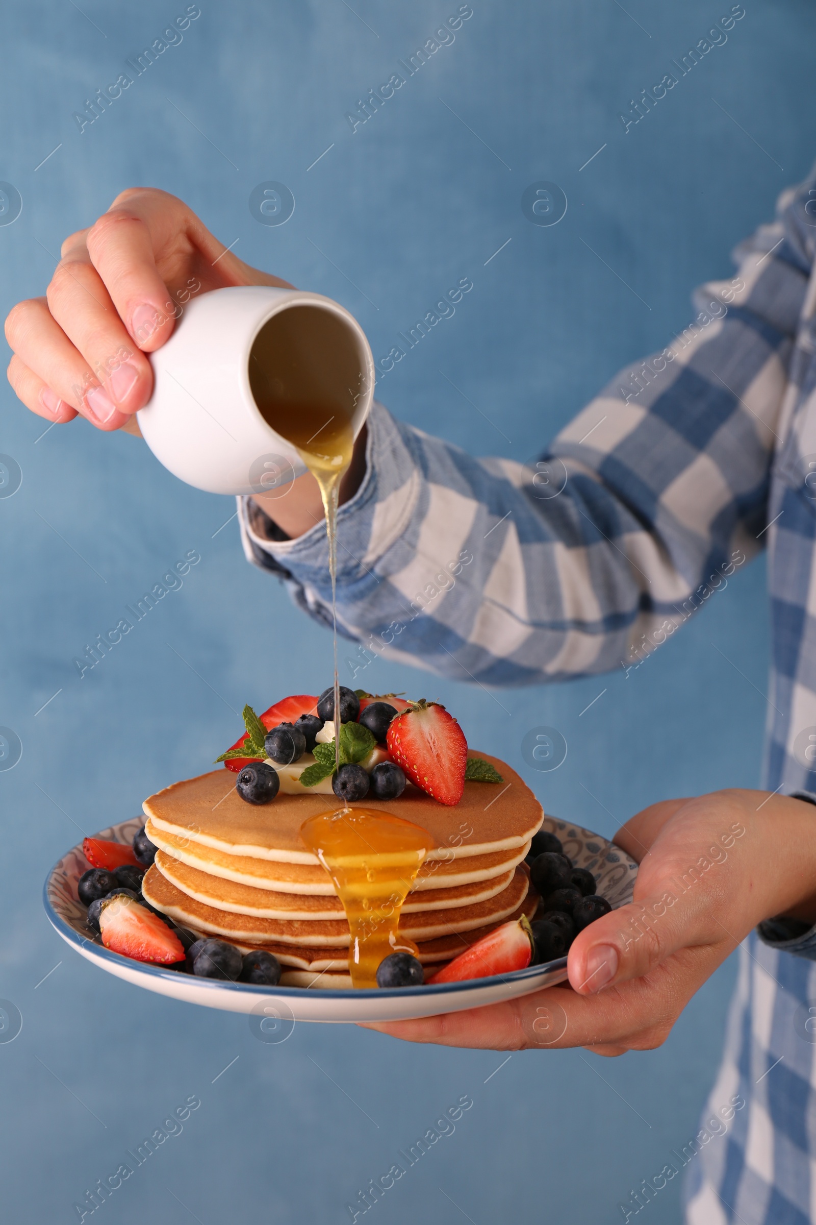 Photo of Woman pouring honey onto delicious pancakes with fresh berries and butter against light blue background, closeup