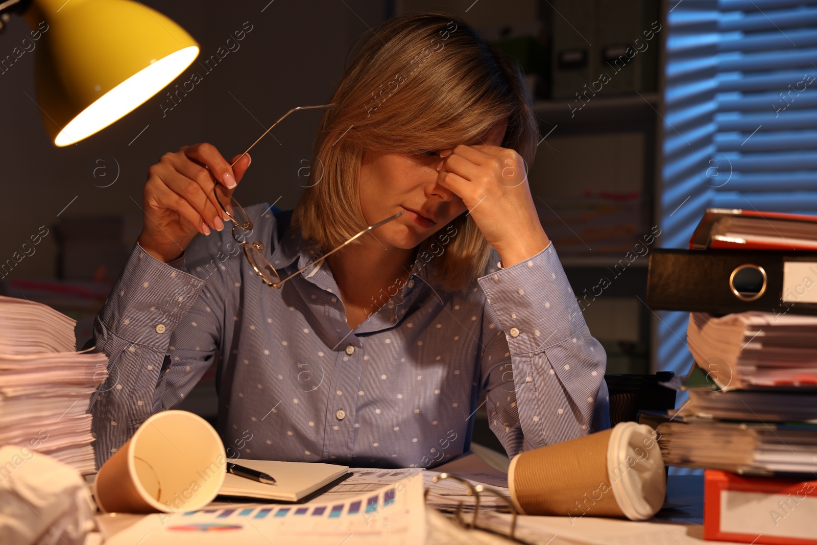 Photo of Overwhelmed woman surrounded by documents and paper coffee cups at table in office at night