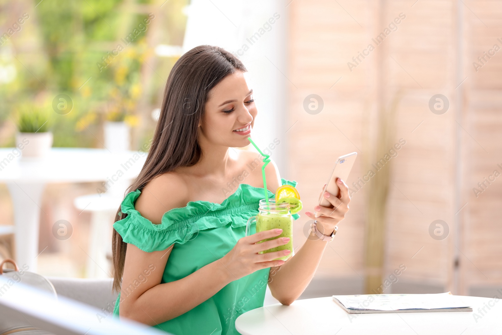 Photo of Young woman using mobile phone while drinking tasty healthy smoothie at table, indoors