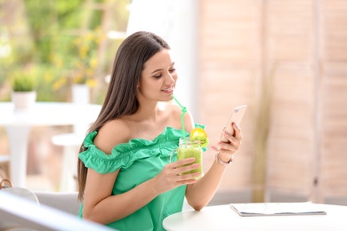 Young woman using mobile phone while drinking tasty healthy smoothie at table, indoors