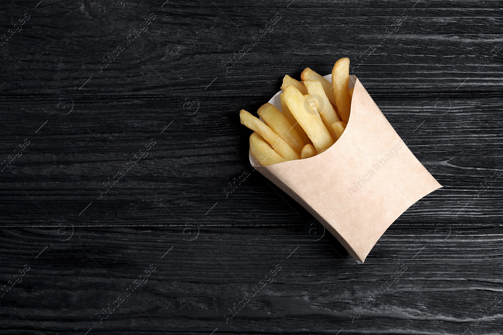 Photo of Delicious french fries in paper box on black wooden table, top view. Space for text